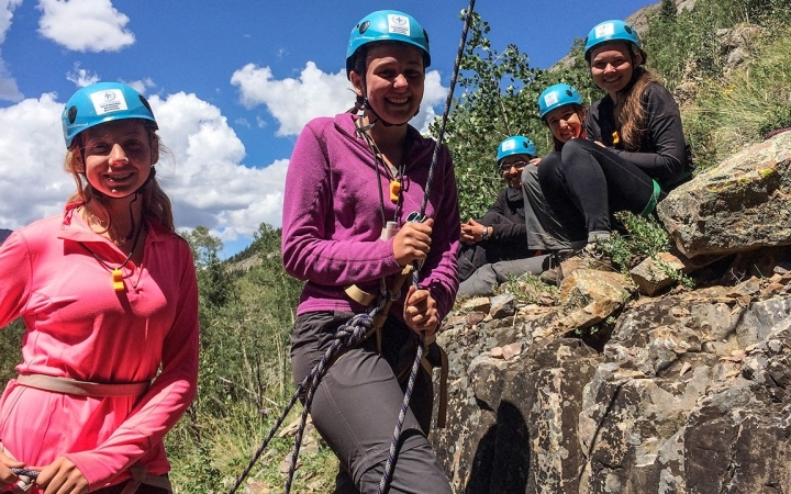 Outward bound students wearing helmets smile at the camera. One person appears to be belaying.